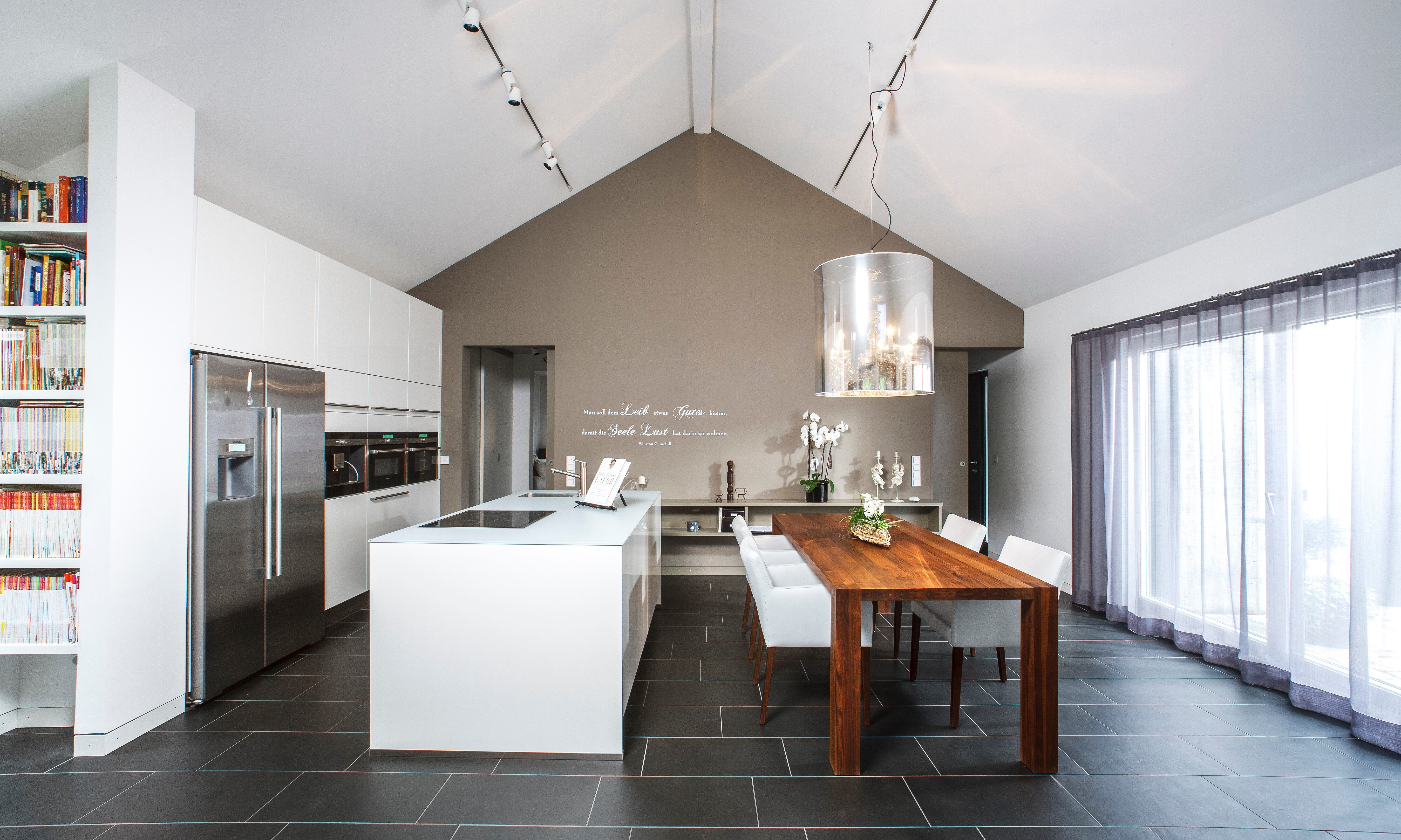 Der massive Esstisch mit weich gepolsterten Stühlen befindet sich unmittelbar neben dem Küchenblock. So kann die Bewohnerin jederzeit mit Gästen kommunizieren. Highlight: Kronleuchter Light Shade Shade. Die bis in den Giebel reichende Wand trennt Wohn- un