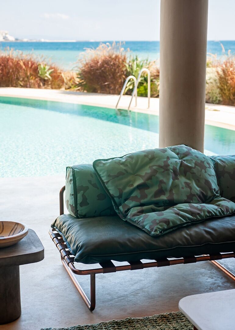 Tagesbett mit grüner Polsterung und Camouflage-Kissen vor Pool und Blick aufs Meer