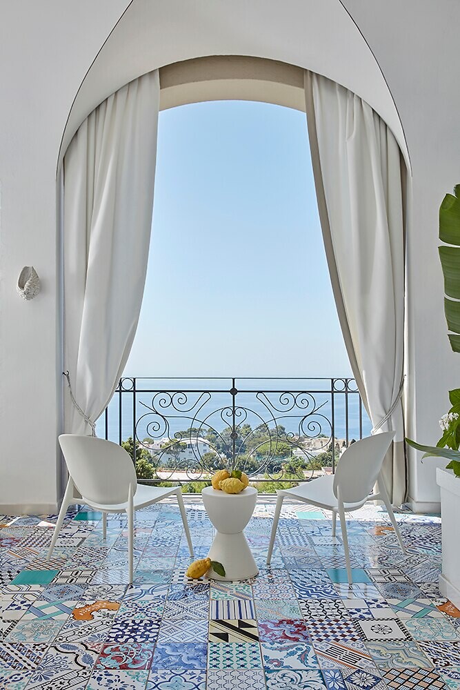 Mediterrane Szene mit Kartell-Stühlen auf farbigem Fliesenmuster Zwei weiße Stühle vor Balkon mit gemustertem Boden und Blick aufs Meer