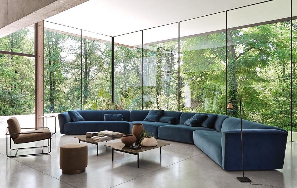 Großzügige Sofalandschaft von Frag in naturnaher Architekturkulisse langes geschwungenes Sofa in Blau in verglastem Wohnbereich mit Blick ins umgebende Grün